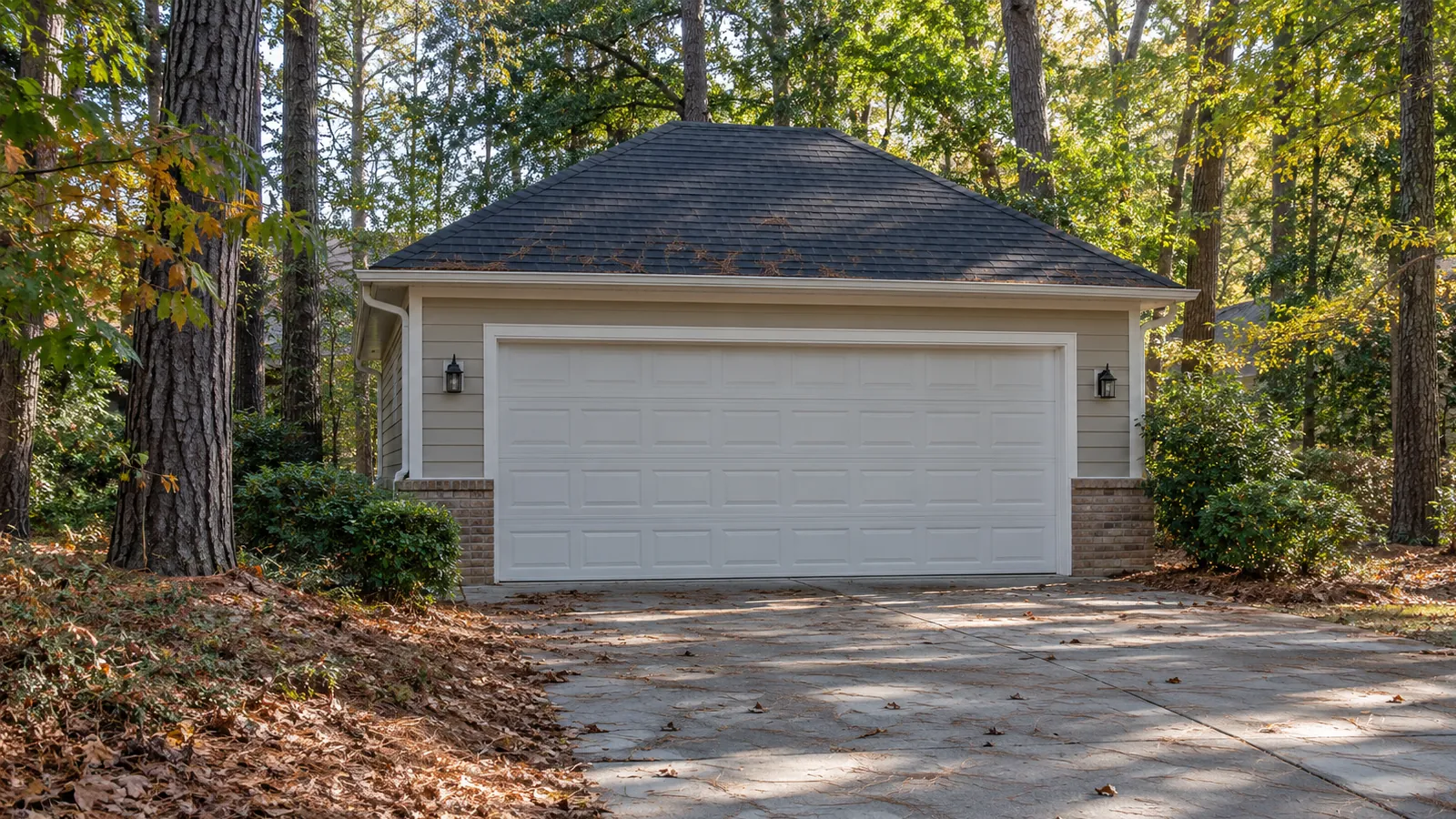 Two car garage with a white raised panel door under tall pines and oaks on a wooded lot, with pine needles along the edge of the driveway