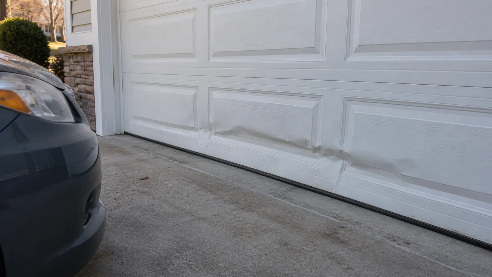 Bottom section of a white steel garage door with a large horizontal dent and crease from a car bumper, seen from the driveway